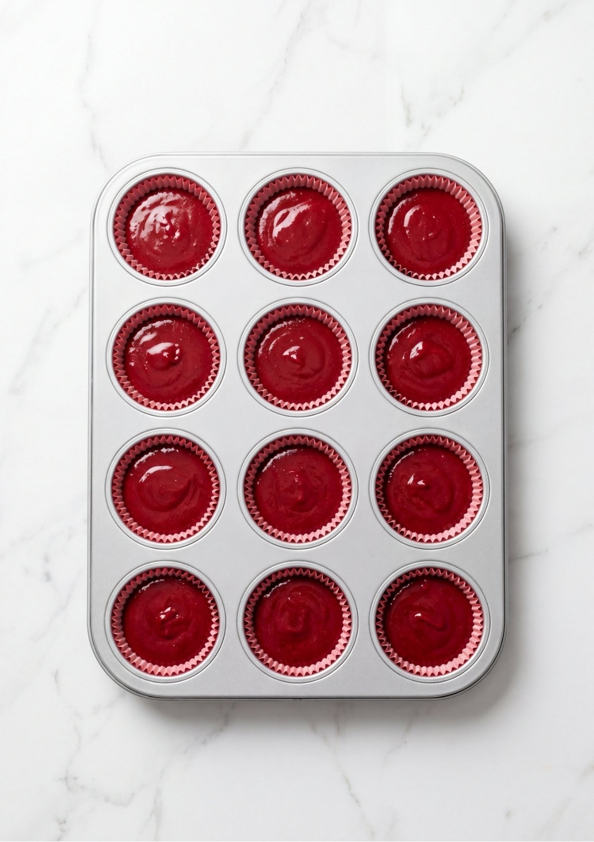 A silver muffin pan filled with smooth, bright red velvet cupcake batter in red paper liners before baking.