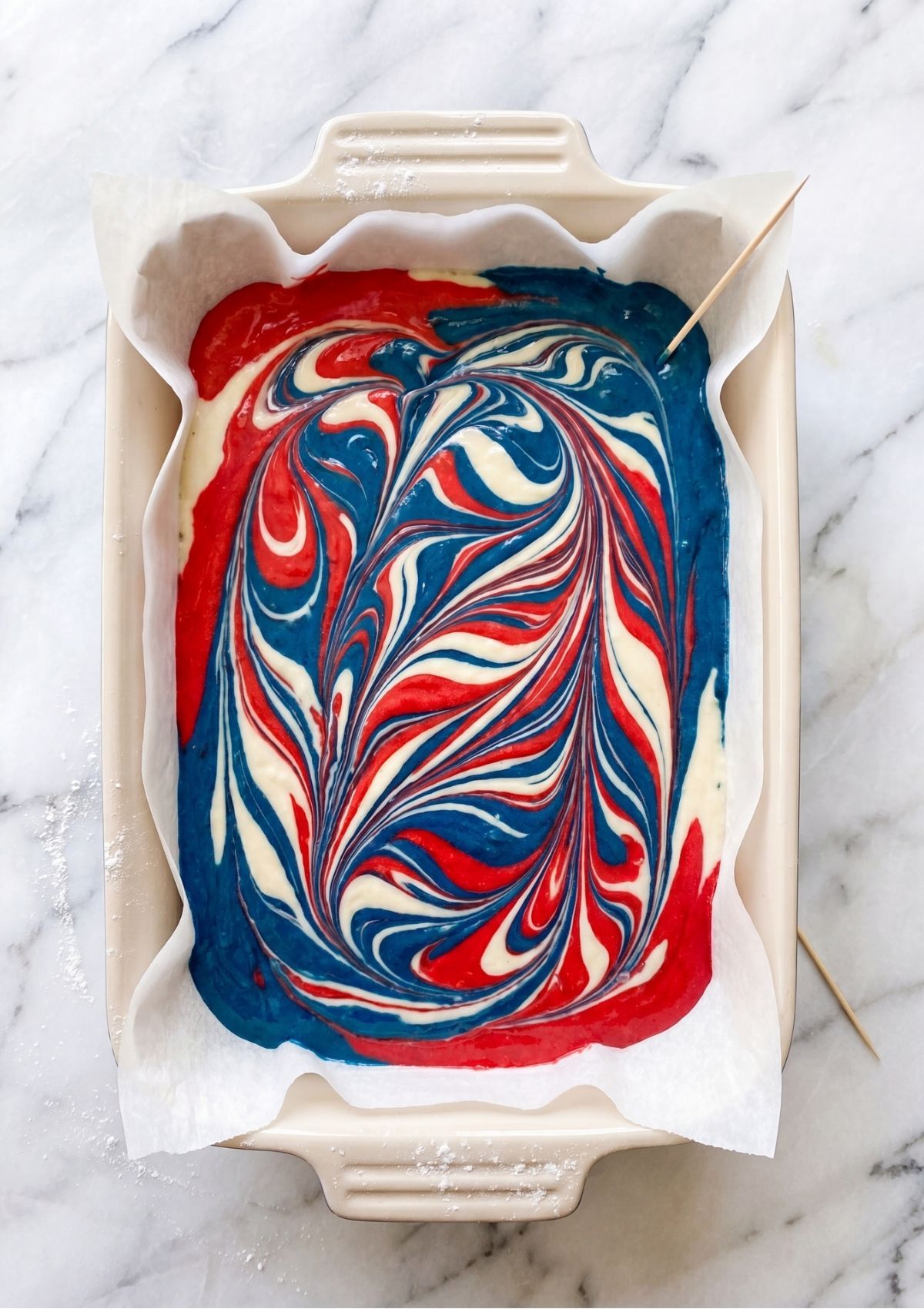 Unbaked red, white, and blue cake batter swirled together in a parchment-lined pan with a toothpick to create a marbled pattern.