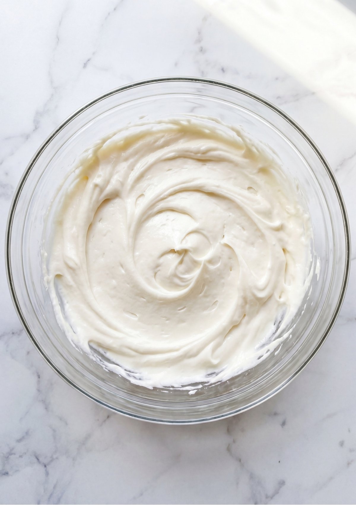 A glass bowl containing thick and fluffy white cream cheese frosting with visible swirl marks.
