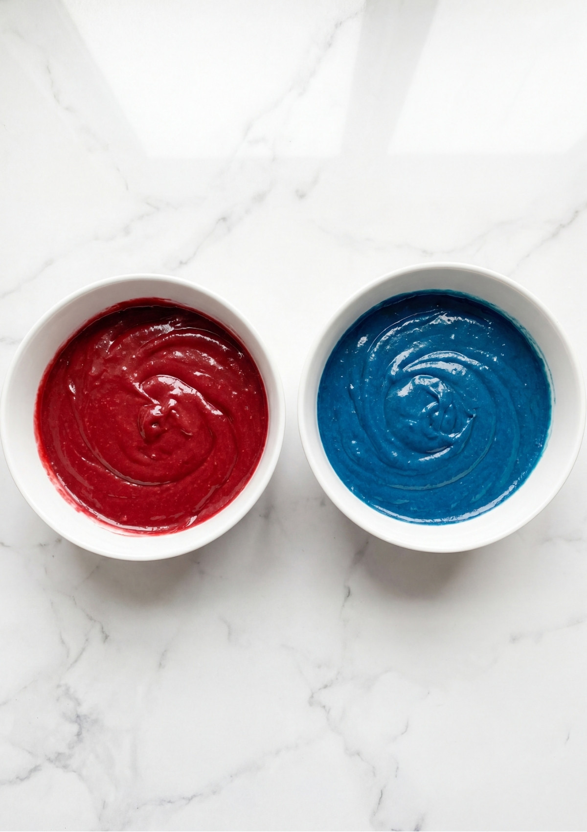 Two white bowls on a marble surface containing bright red and deep blue cake batter.