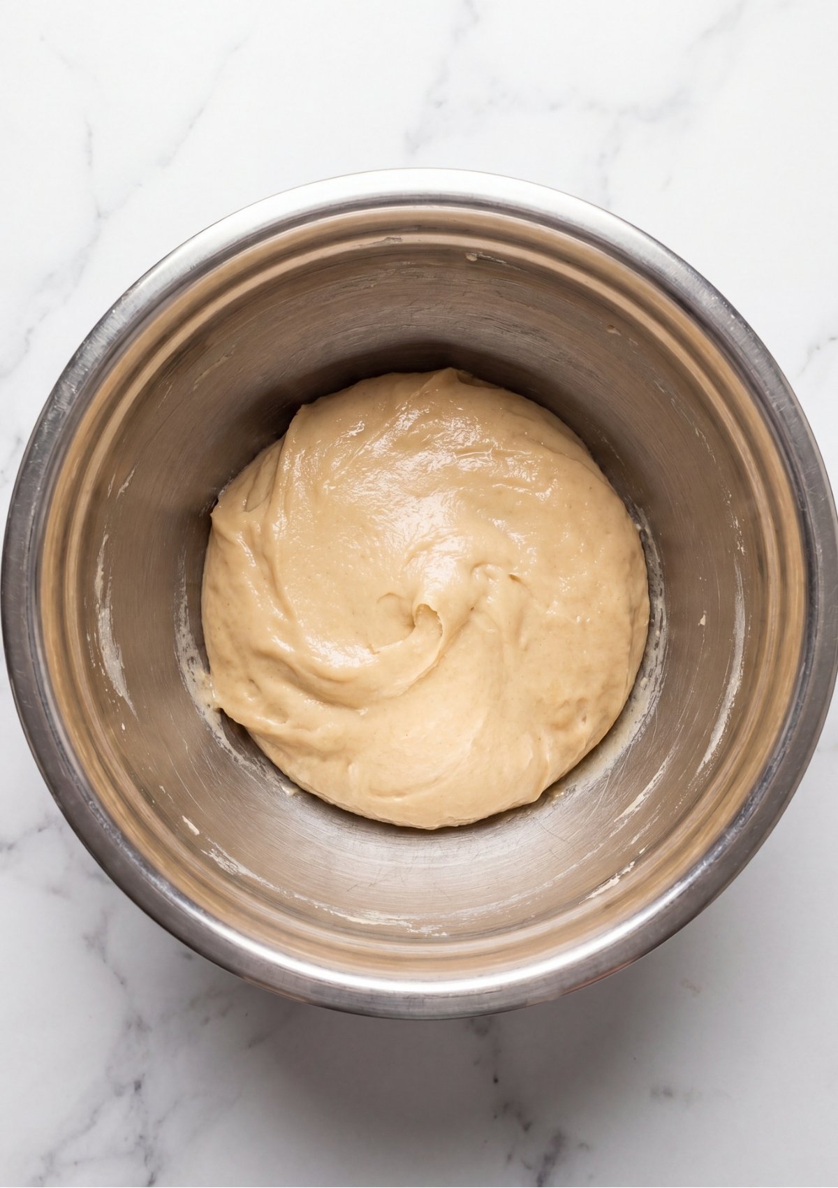 A smooth, well blended pale tan dough base inside a stainless steel mixing bowl on a marble background
