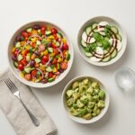A spread of low-sugar, vegetable-forward dishes including an Italian chopped salad, a cucumber caprese salad, and a healthy avocado potato salad.
