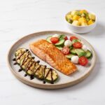 A plate with pan-seared salmon, balsamic grilled zucchini, cucumber caprese salad, and a side bowl of tropical fruit salad.
