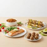 A spread of protein-forward dishes including pan-seared salmon, lemon pesto chicken kabobs, high-protein snack balls, and roasted chickpeas.