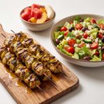 Lemon pesto chicken kabobs on a wooden board alongside a bowl of chopped salad and fruit.
